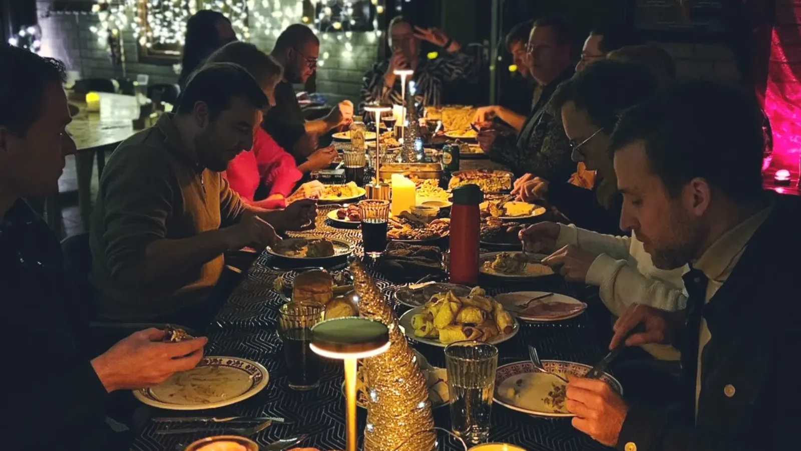 Spelers schuiven aan voor het gourmetdiner aan een lange tafel vol schalen, bij kaarslicht en kerstverlichting